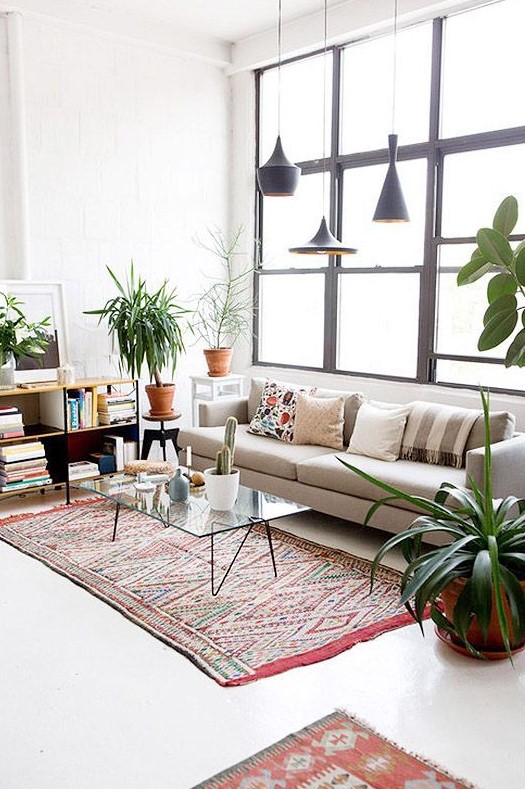 a modern boho living room with a neutral cotton sofa, printed rugs, a bookshelf, a glass coffee table, black pendant lamps and potted plants