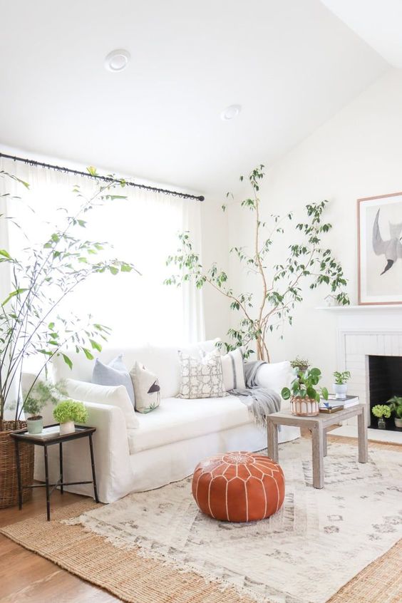 a chic neutral living room with a fireplace, a white cotton sofa by IKEA, a wooden table, an amber leather pouf and greenery