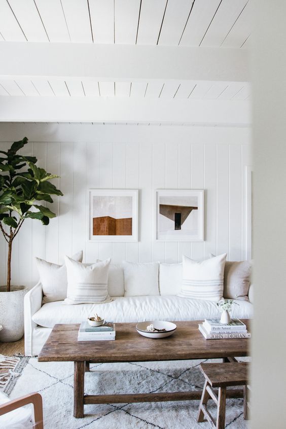 a neutral living room with a white cotton sofa and pillows, a stained coffee table and a stool, a potted tree and a mini gallery wall