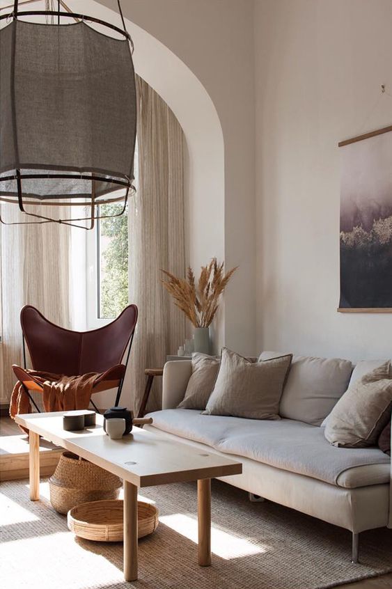 a boho living room with a neutral olefin sofa, a wooden table, a burgundy butterfly chair, a pendant lamp and some art