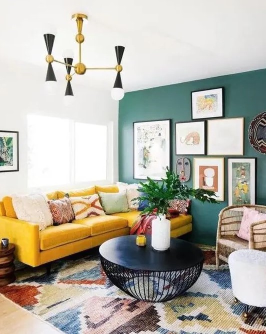 a bright boho living room with a green wall and a gallery wall, a yellow velvet sectional, a retro chandelier and a black round table