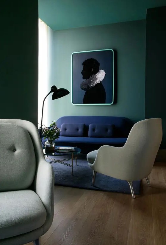 a chic and stylish living room with green walls, a navy loveseat, grey chairs, a glass coffee table and a bold and contrasting artwork