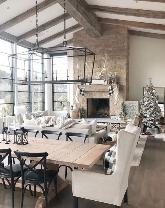 a barn living room with a glazed wall, a fireplace clad with stone, wooden beams on the ceiling, neutral seating furniture and pendant lamps