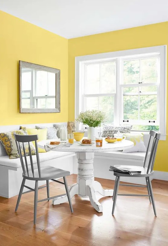 a colorful dining nook with yellow walls, a white corner seat, a white table, grey chairs and grey and yellow pillows