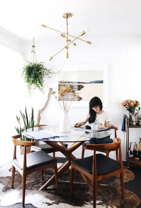 a cool modern farmhouse dining room with a glass top dining table, rich-stained wishbone chairs with black seats, a cowhide rug and greenery