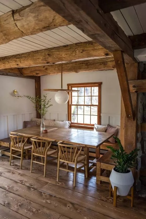 a cozy rustic dining space with stained wooden beams, a built-in bench, a stained table and chairs, a lovely pendant lamp and greenery