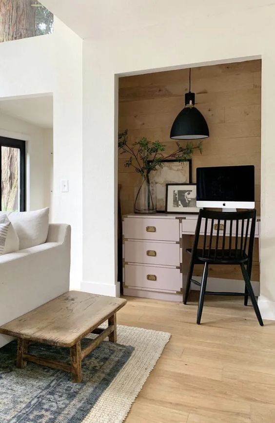 a deep niche clad with wood, with a pink desk, a black chair, a pendant lamp and some artwork and greenery