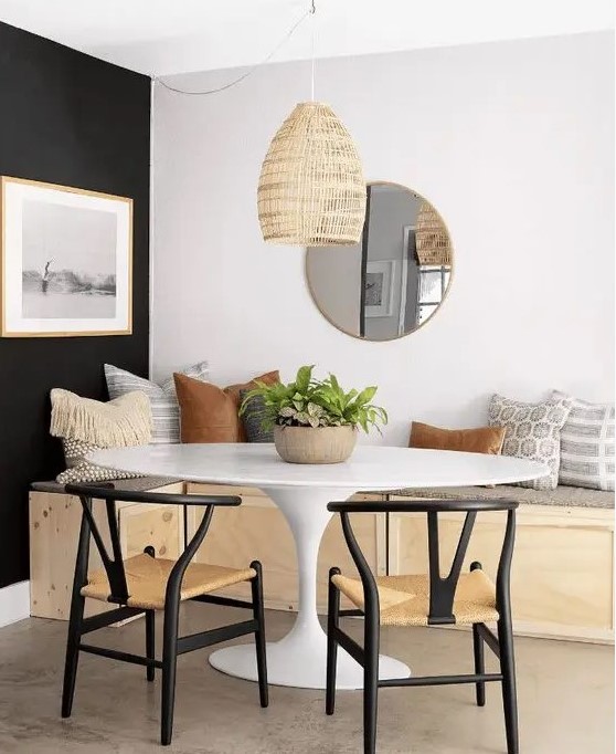 a farmhouse dining room with a built-in bench with pillows, an oval table and black wishbone chairs and a round mirror