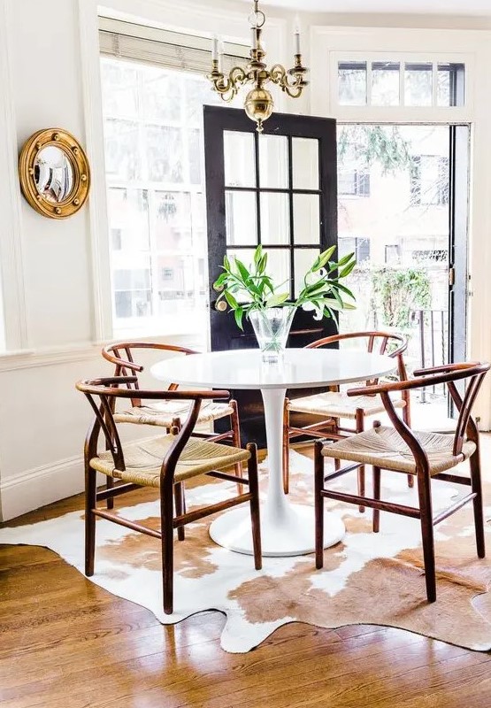 a farmhouse dining room with a round table, rich-stained wishbone chairs, a cowhide rug, a gilded chandelier