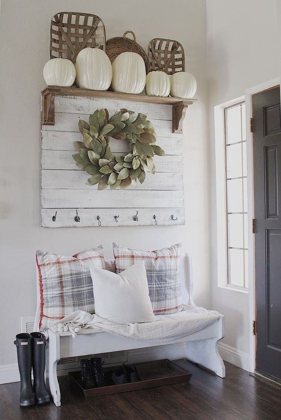 a farmhouse entryway with a white bench, checked pillows, a white rack, a wreath and a rack with white pumpkins for fall