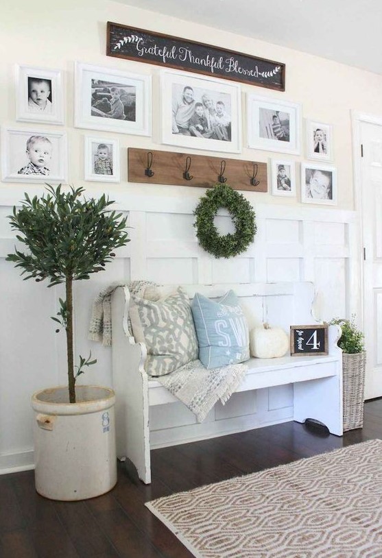 a farmhouse entryway with a white bench with pillows, a pumpkin, a gallery wall, a greenery wreath and a potted tree