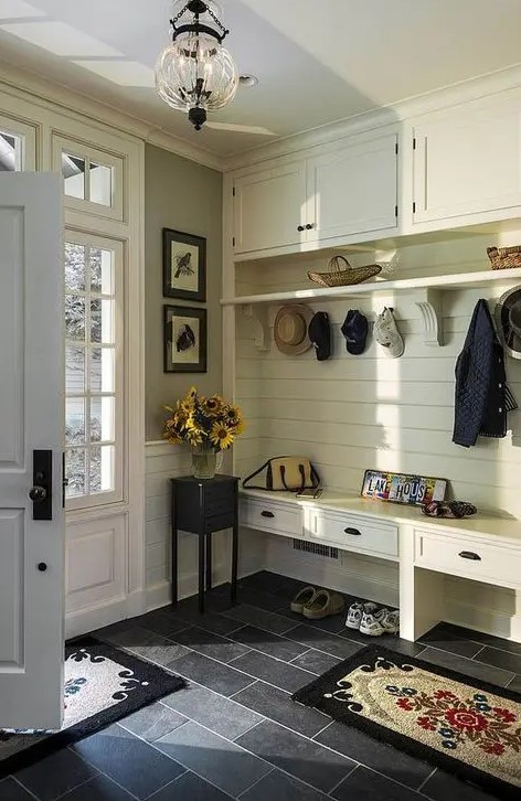 a farmhouse entryway with an ivory wooden furniture piece iwth a built-in bench of drawers and more storage space plus dark tiles on the floor for a contrast