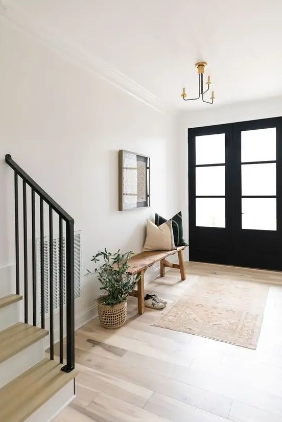 a farmhouse entryway with black doors, a stained bench and black and white pillows, a printed rug and a potted plant