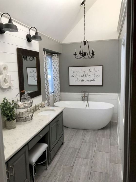 a grey farmhouse bathroom with a large vintage vanity, a stone countertop, an oval tub, a chandelier and some vintage wall lamps