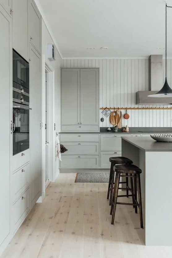 a light grey modern country kitchen with shaker style cabinetry, black countertops and built-in appliances, a modern hood and tall stools