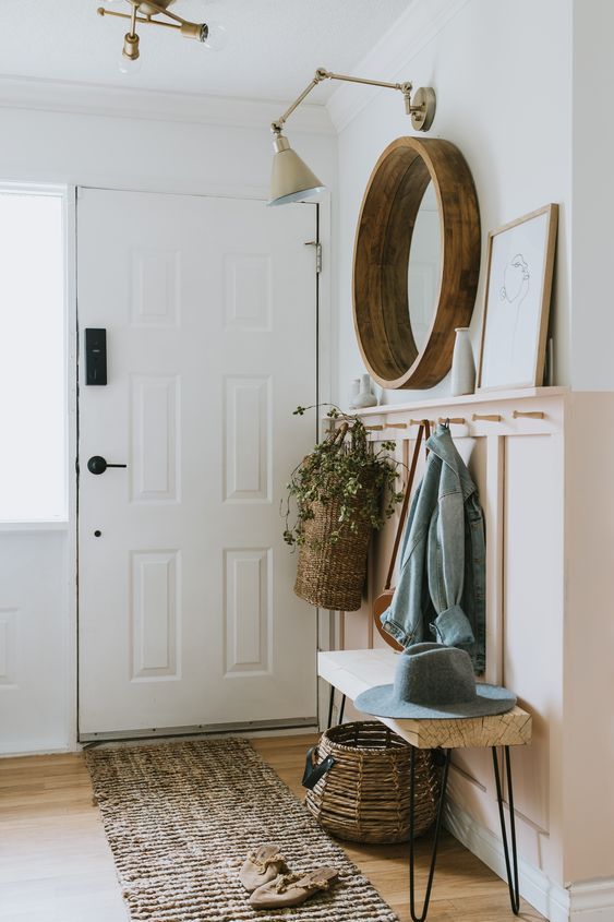 a lovely modern farmhouse entryway with a pink panaled wall with books, a hairpin leg bench, a mirror in a round frame and a sconce