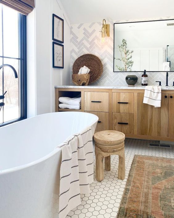 a modern country bathroom with herringbone and hex tiles, an oval tub, a timber vanity, a wooden stool, a mirror and some decor