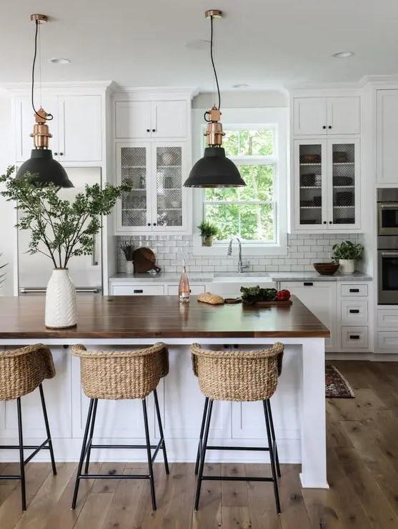 a modern country kitchen in white, with cool cabinets, a large kitchen island with a butcherblock countertop, pendant lamps and woven stools