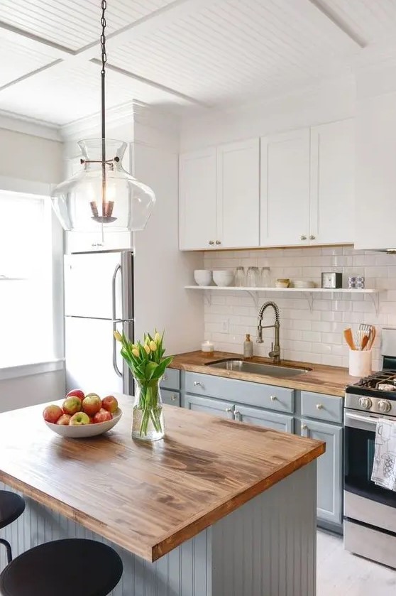 a modern country kitchen with white and light grey cabinets, butcherblock countertops, a shiny subway tile backsplash and a pendant lamp