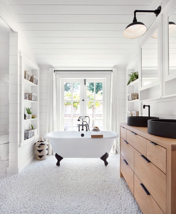 a modern farmhouse bathroom with a pebble floor, shiplap, a timber vanity, black sinks and a clawfoot tub by the window