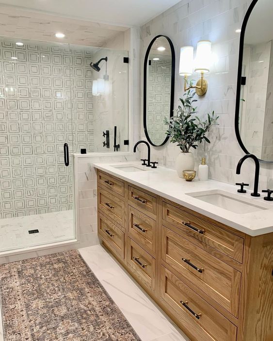 a modern farmhouse bathroom with printed and marble tile, a stained vanity, oval mirrors, black hardware and a boho rug