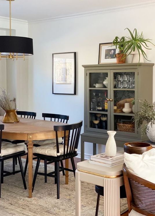 a modern farmhouse dining room with an olive green cupboard, an oval stained table, black farmhouse chairs, a black chandelier and some art and decor