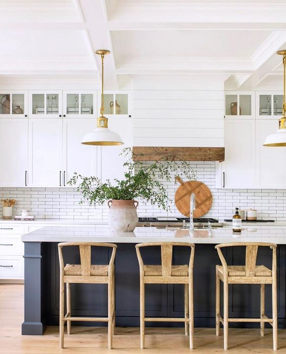 a modern farmhouse kitchen with white cabinets, a soot kitchen island, pendant lamps, woven stools and touches of wood