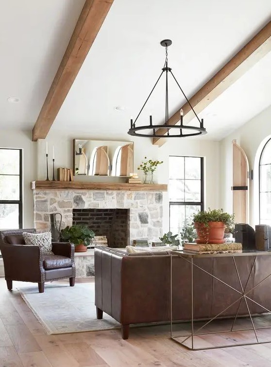 a modern farmhouse living room with a stone clad fireplace, wooden beams, leather furniture, a metal chandelier and potted plants