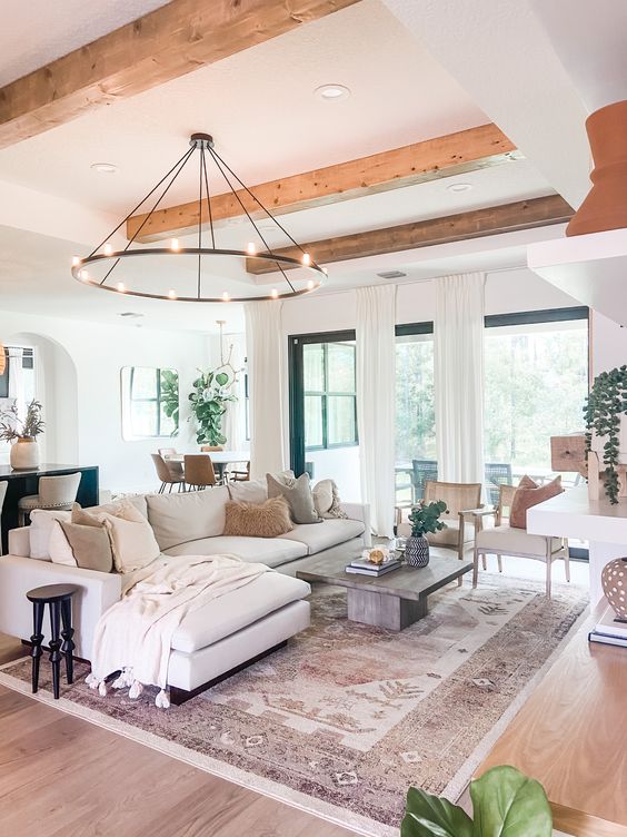 a modern farmhouse living room with wooden beams, a chandelier, a neutral sectional, a coffee table, a fireplace, some chairs