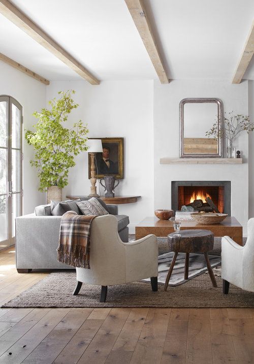 a modern farmhouse living room with wooden beams, a fireplace, a grey sofa, neutral chairs, a coffee table and some greenery