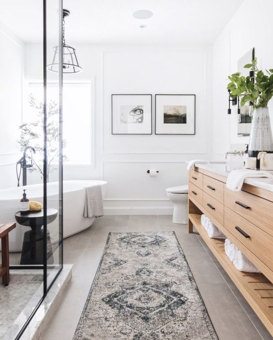 a modern farmhouse meets boho bathroom with a tub by the window, a large timber vanity, a shower space, a boho rug and some greenery