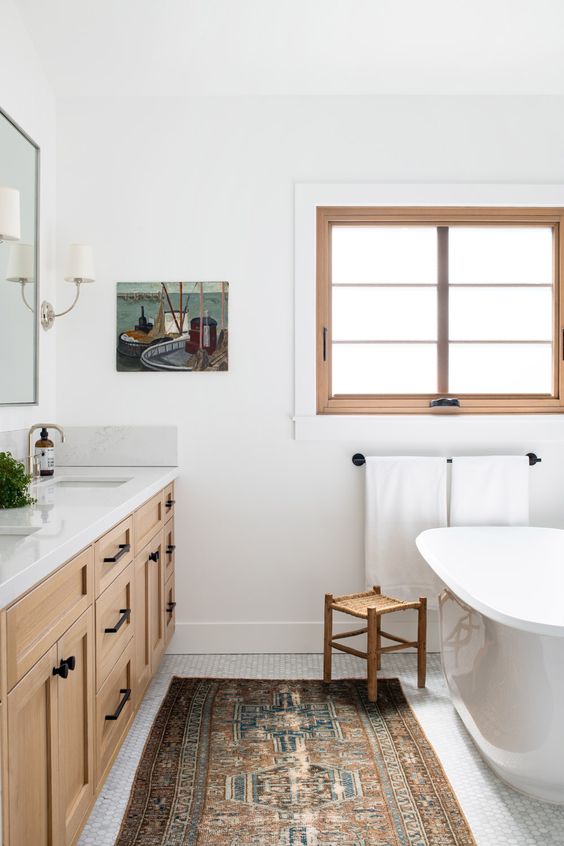 a modern farmhouse meets boho bathroom with a window, a timber vanity, a tub, a stool, a boho rug and some lovely decor