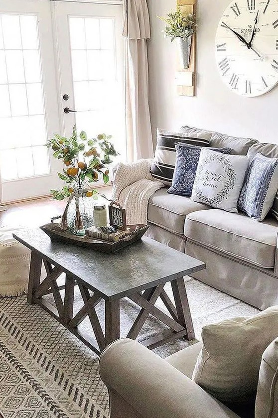 a neutral farmhouse living room with grey furniture, a wood and concrete coffee table, a large clock and printed textiles