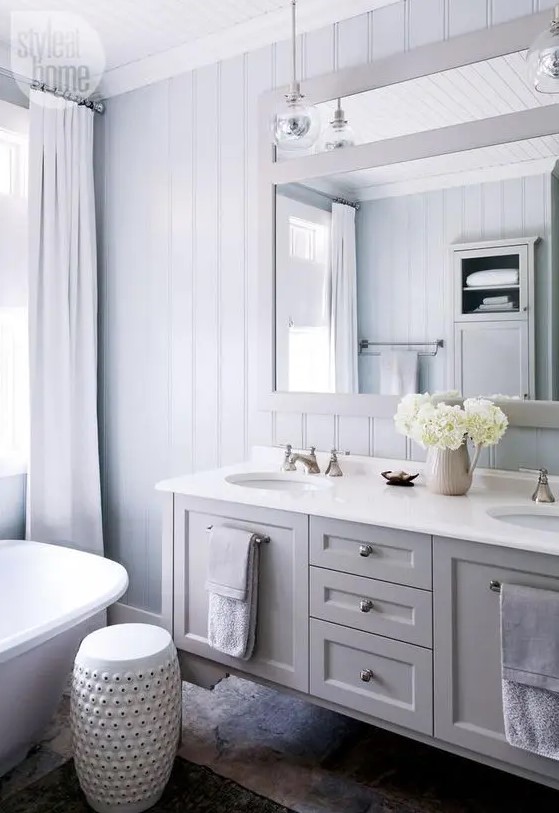 a neutral modern country bathroom with pale blue planked walls, a floating grey vanity, a large mirror, pendant lamps and neutral textiles