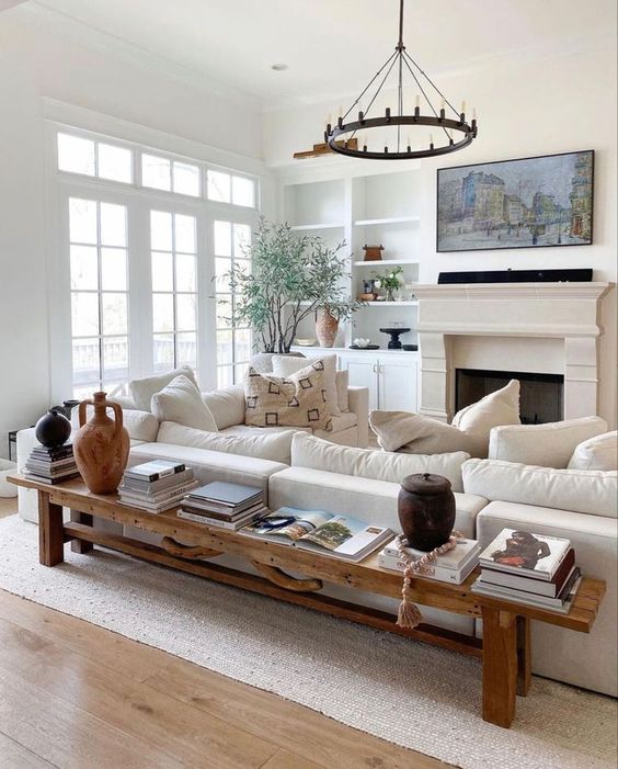 a neutral modern farmhouse living room with built-in shelves, a fireplace, a creamy sofa, a bench as a coffee table and a chandelier