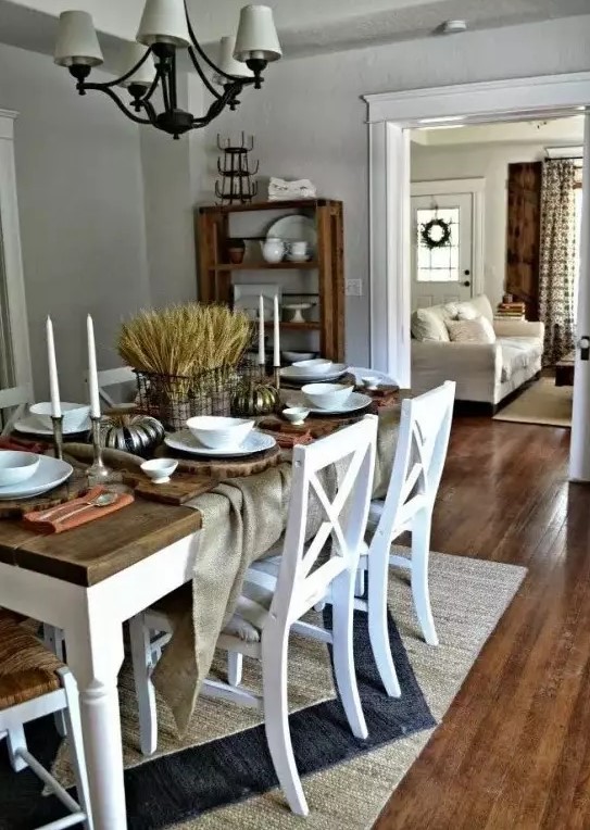 a rustic vintage dining space with grey walls, a white table with a stained tabletop, white chairs, a vintage chandelier and wheat centerpiece plus some candles