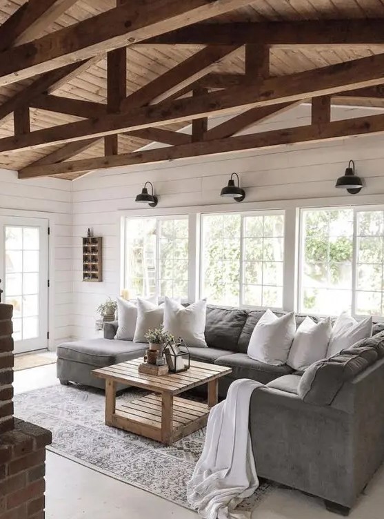 a simple barn living room with white planked walls, a grey sofa, wooden beams, a wooden table and lots of natural light