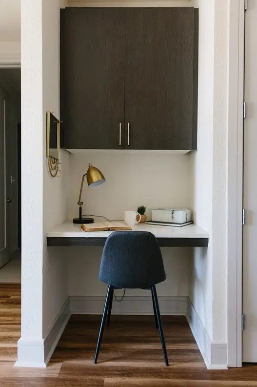 a tiny home office built into a small nook, with a built-in desk and a large cabinet, a navy chair, a brass table lamp