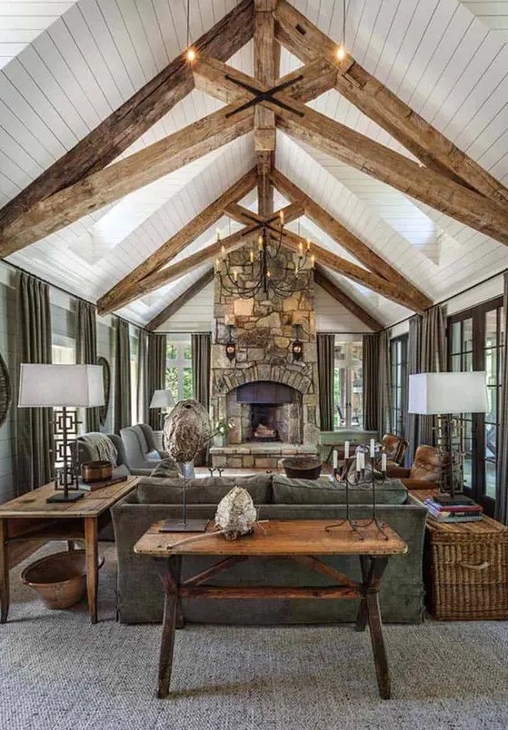 a welcoming barn living room with wooden beams, a fireplace clad with stone, grey seating furniture, a metal chandelier and baskets