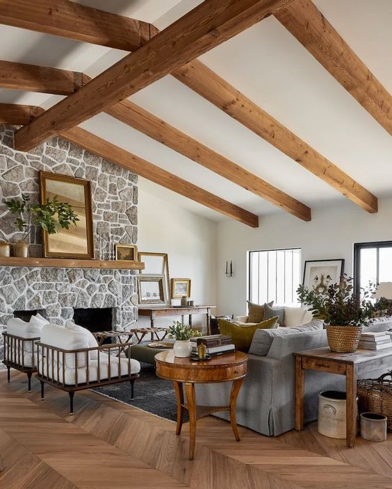 a welcoming modern farmhouse living room with wooden beams, a fireplace clad with stone, a grey sofa and white chairs, stained and refined furniture