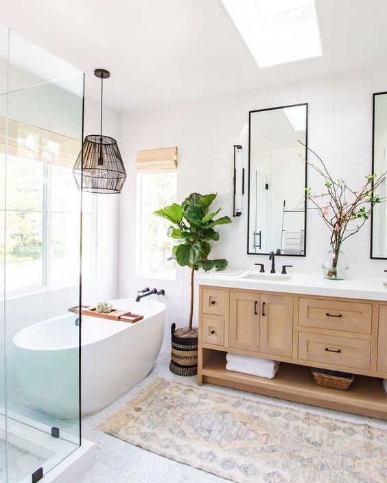 an airy modern farmhouse bathroom with a double timber vanity, framed mirrors, an oval tub, a boho rug, a black pendant lamp and a boho rug