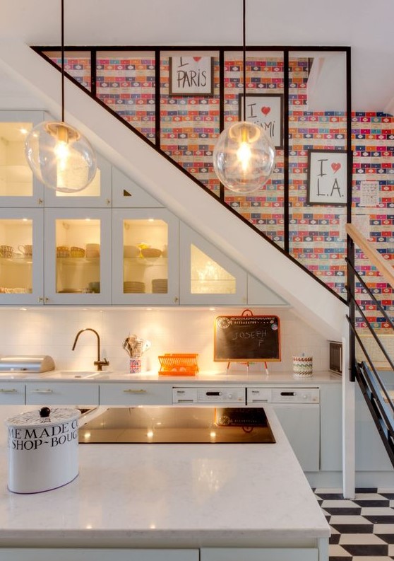 a catchy modern kitchen built into the space under the stairs, with built-in lights and glass doors, a kitchen island and pendant lamps