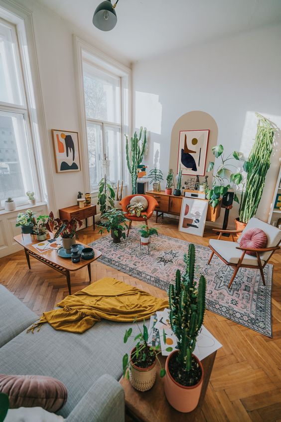 a colorful boho living room with a grey sectional, a printed rug, some chairs, a lot of potted plants and artwork