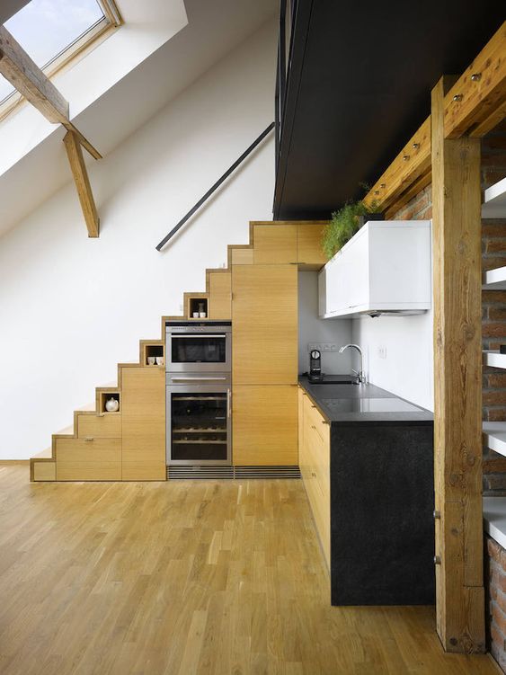 a modern kitchen with stained cabinets built into the stairs, black countertops, upper white cabinets and greenery