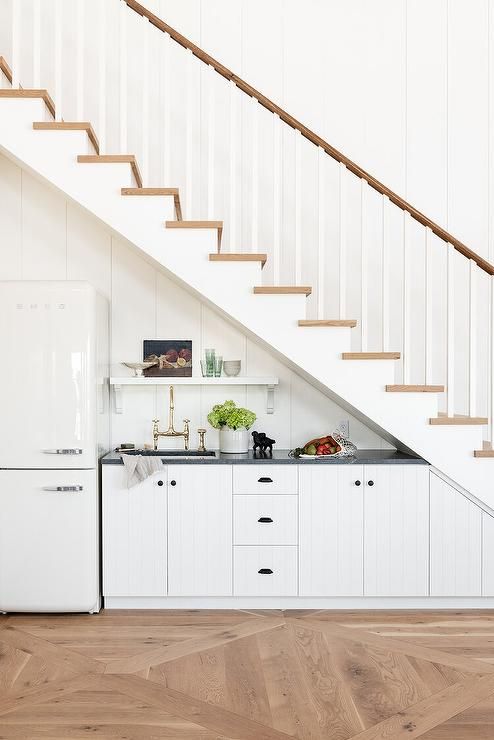 a pretty farmhouse under the stairs kitchenette with planked cabients, black countertops, a fridge and a shelf is cool