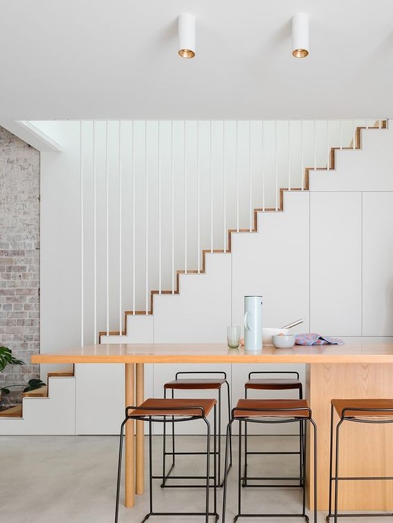 a sleek minimalist kitchen with a staircase that hides storage compartments, a kitchen island, tall stools and spotlights