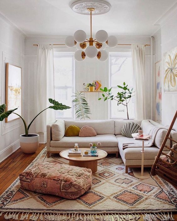 a small boho living room with a white sectional, a printed boho rug and a pouf, some bright art, pillows and a chandelier