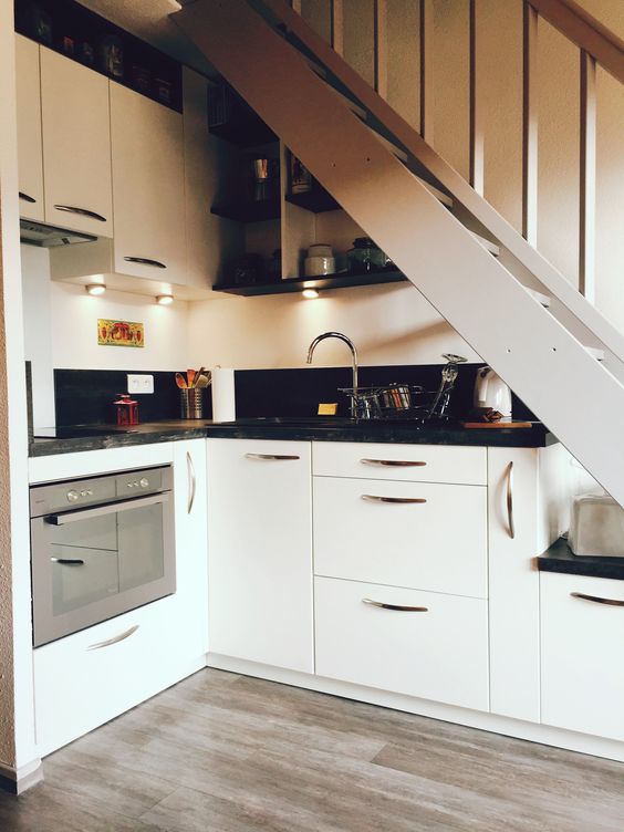 a small kitchen built under the staircase, with white cabinets and black countertops, built-in lights is a lovely space