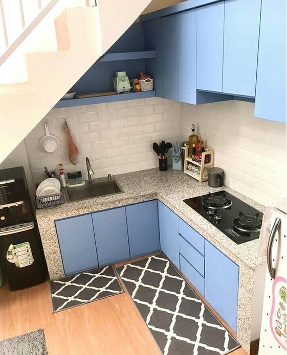 a tiny blue kitchen built under the staircase, with a subway tile backsplash and terrazzo countertops and black rugs