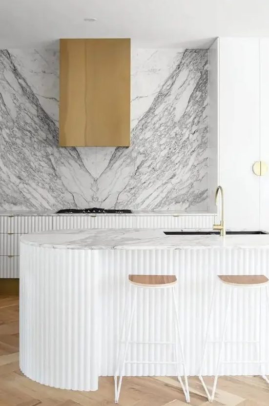 40 A Refined Contemporary Kitchen With Ridged Cabinets And A Matching Curved Kitchen Island A White Marble Backsplash Plus A Brass Hood 